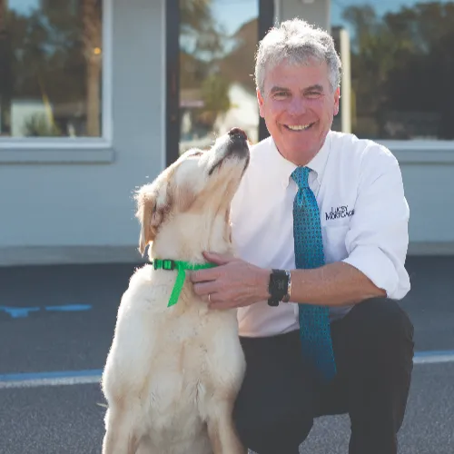 Lorcan Lucey, smiling in a Lucey Mortgage shirt and teal tie, kneeling beside a golden retriever